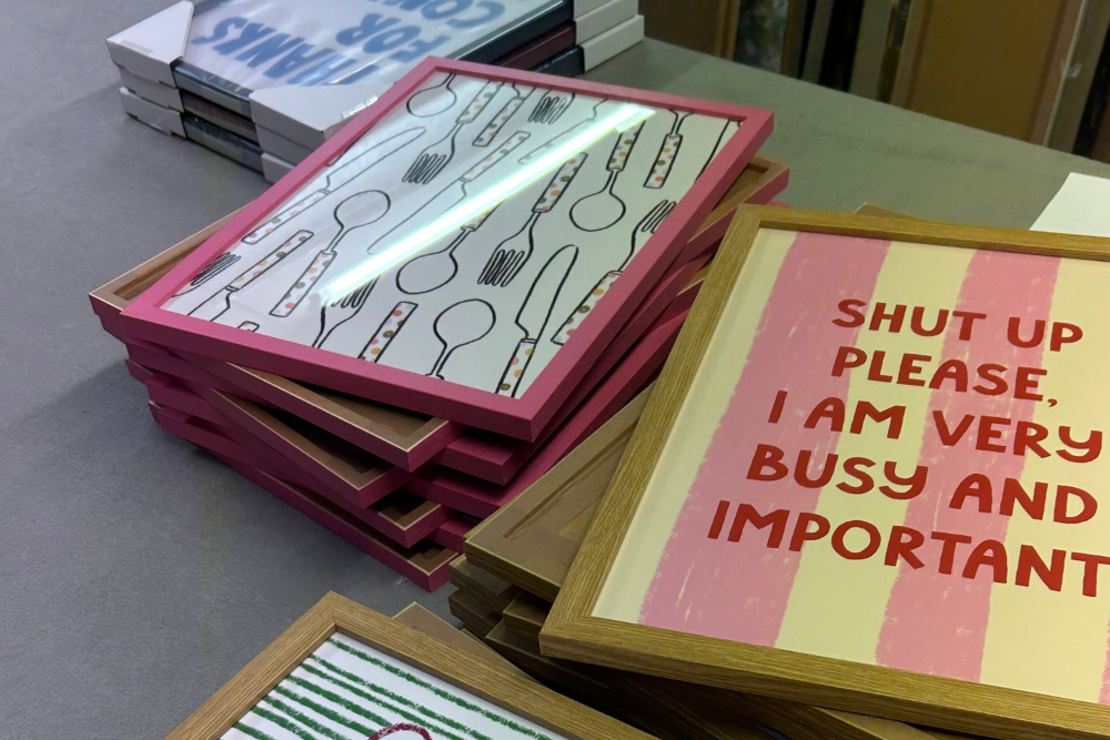 Stacks of framed art prints on a gray table, including one with pink frames and utensil illustrations, another with red text on pink and cream stripes, and another with green stripes at the bottom.